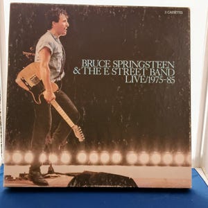 May include: A square box set for Bruce Springsteen & The E Street Band Live/1975-85 featuring 3 cassettes. The cover shows a musician in a light shirt and dark pants playing a light-colored electric guitar on a stage with bright stage lights.
