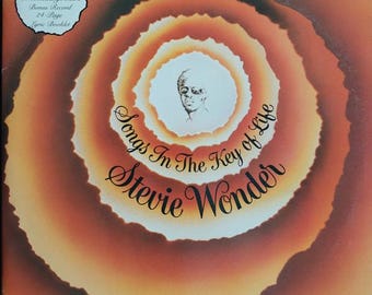 May include: A vintage vinyl record album cover featuring a swirling orange and yellow background with a black and white illustration of Stevie Wonder in the center. The album title "Songs In The Key of Life" is written in white cursive lettering.