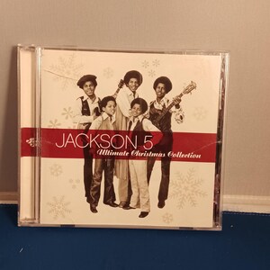 May include: A Jackson 5 Ultimate Christmas Collection music CD in a clear plastic jewel case. The cover features a black and white photograph of the five band members, a red horizontal stripe, and white snowflake patterns on a cream background.