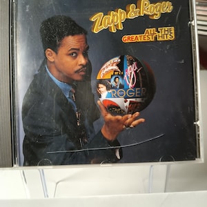 May include: CD case for the album "Zapp & Roger: All the Greatest Hits." The cover features a man in a suit holding a globe-like image with the word "Roger" in a stylized font. The album title is in gold lettering.