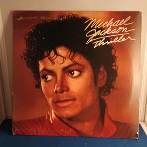 May include: A square vinyl record cover featuring a portrait of Michael Jackson with dark curly hair, wearing a red jacket. The text reads Michael Jackson Thriller and Special 12" Dance Single in white and gold script against a warm brown background.