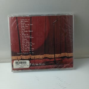 May include: A CD case with a red stage curtain design and a microphone stand. The tracklist includes songs such as "White America" and "Superman." The CD is sealed in plastic.