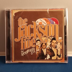 May include: A CD case featuring The Jackson Five The Steeltown 45's album cover. The brown cover displays the group's name in large orange lettering, four small circular record labels, and a vertical strip of black and white photographs of the band members.