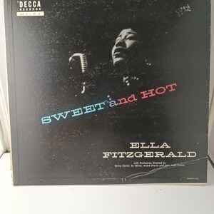 以下が含まれることがあります： エラ・フィッツジェラルドの「Ella: Sweet and Hot」のヴィンテージビニールレコードアルバムカバー。カバーには、マイクを持ったフィッツジェラルドの白黒画像と、青と赤のテキストでタイトルが描かれています。