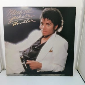 May include: Album cover of Michael Jackson in a white suit and black shirt. The album title "Thriller" is written in gold script. The background is a dark gradient.