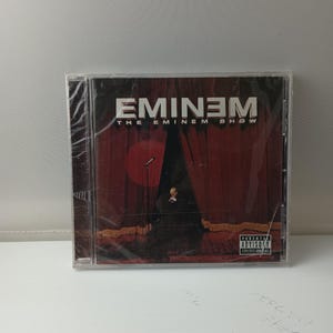 Puede incluir: Se muestra la carátula del CD del álbum de Eminem, "The Eminem Show". La portada del álbum presenta un escenario con cortinas rojas, un micrófono y una figura con traje. El título del álbum y el nombre del artista están en texto blanco sobre un fondo oscuro.
