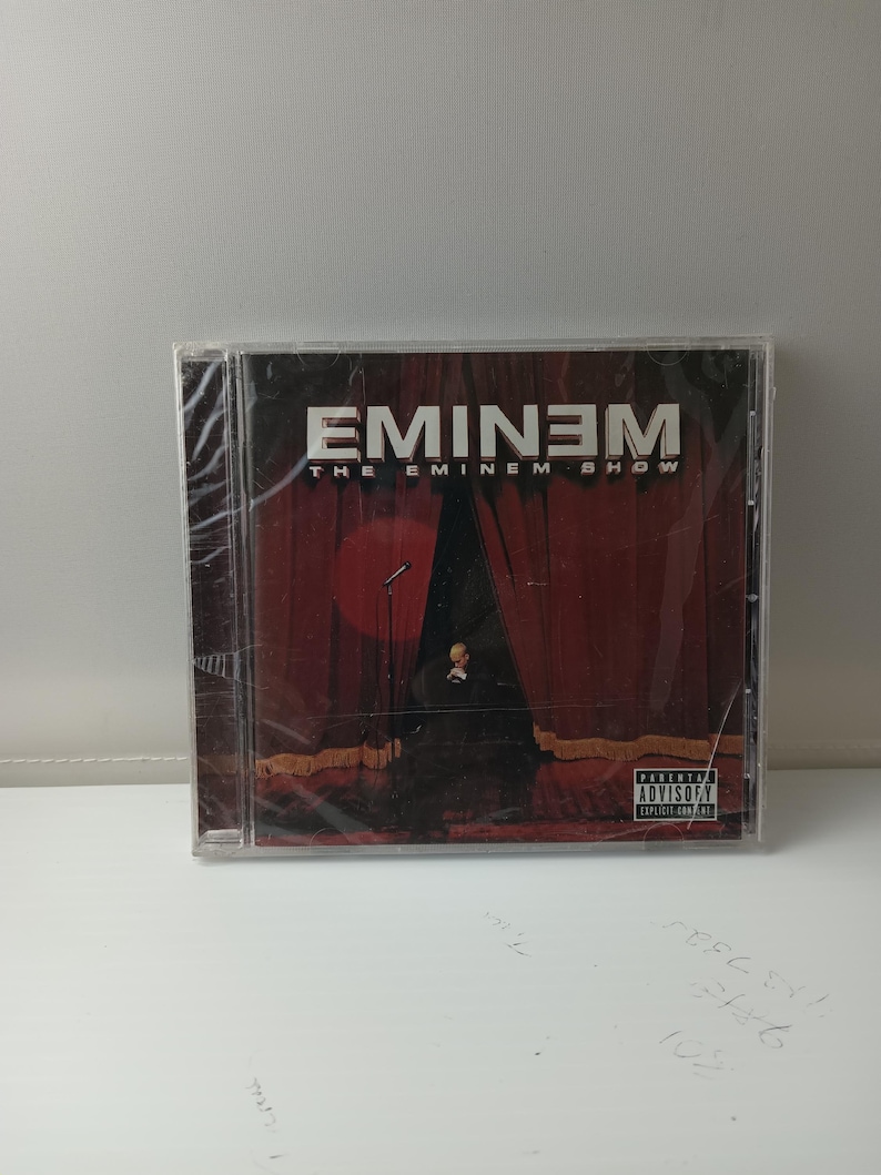 May include: A CD of Eminem's album, "The Eminem Show," is displayed. The cover art depicts a stage with red curtains, a microphone, and a figure in a suit. The album title and artist name are in white text against a dark backdrop.