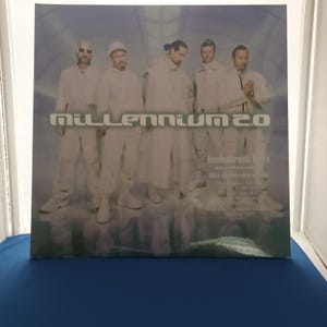 Può includere: Una copertina quadrata per disco in vinile che mostra cinque persone con abiti bianchi coordinati in un ambiente futuristico azzurro. Il testo "MILLENNIUM 2.0" è stampato con un carattere stilizzato al centro della copertina dell'album.