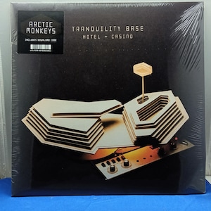 Peut inclure: Une pochette de disque vinyle pour Arctic Monkeys intitulée Tranquility Base Hotel + Casino. La couverture noire présente une structure de modèle architectural beige en couches et un petit panneau de contrôle avec des boutons.
