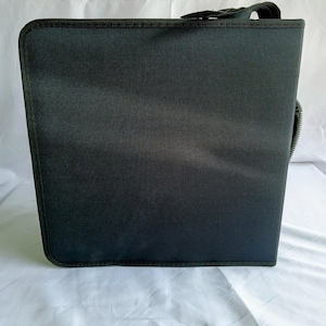 May include: A square black storage case with a textured fabric exterior and reinforced stitched edges. The case features a black handle attached to the top and a visible silver zipper closure along the side, providing a secure enclosure for contents.