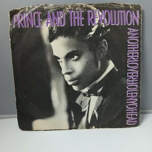 May include: A vintage album cover for Prince and The Revolution, featuring a black and white portrait of Prince. The album title, "Anotherloverholenyohead," is printed in purple, along with the band's name. The cover has visible wear.
