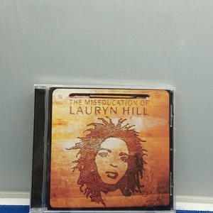 May include: A compact disc case featuring the album cover for The Miseducation of Lauryn Hill. The artwork displays a stylized portrait of a person with textured hair against a warm, orange and brown gradient background with the title text.