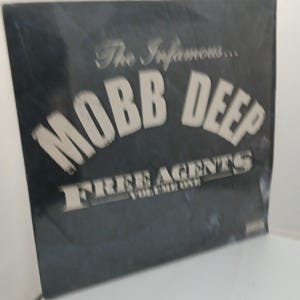 May include: A black album cover with the text "The Infamous... MOBB DEEP FREE AGENTS VOLUME ONE" in white lettering. The album cover is slightly angled, showcasing the title and artist.