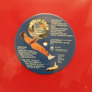 Può includere: Un disco in vinile rosso dei Jonzun Crew, con Michael Jonzun. L'etichetta blu mostra titoli di canzoni come "Redd Hott Mama" e dettagli di produzione. È una copia promozionale.