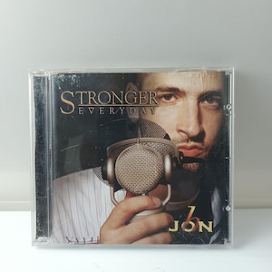 May include: CD case featuring a man holding a vintage microphone. The album cover reads "STRONGER EVERYDAY" with the artist's name "JON" in a bold font. The image has a close-up shot of the man's face and hand.