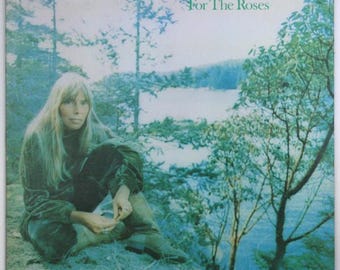 May include: A vintage vinyl record album cover featuring Joni Mitchell. The cover art shows Joni Mitchell sitting on a rock by a lake, with trees and water in the background. The album title "For The Roses" is printed in white text on a blue background.