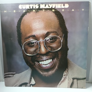 May include: Album cover for Curtis Mayfield's "Heartbeat." The album art features a smiling man wearing glasses. The album title and artist's name are printed in a gold font against a dark background.