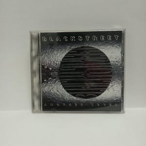 May include: A compact disc case featuring the album "Another Level" by Blackstreet. The cover art is primarily black and silver, with the band's name at the top and album title at the bottom.