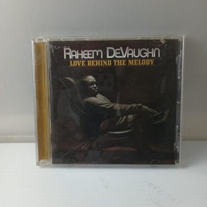 Può includere: Una custodia per CD con l'album "Love Behind the Melody" di Raheem DeVaughn. La copertina mostra un uomo seduto su una sedia. Il testo "Raheem DeVaughn" e "Love Behind the Melody" è visualizzato in bianco e oro.