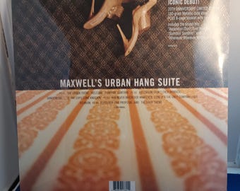 May include: A square vinyl record jacket for Maxwell's Urban Hang Suite. The cover features a pair of tan high-heeled shoes on a dark patterned surface above the title. The lower section shows a blurred orange and white striped pattern with text.