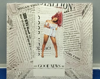 May include: A rectangular display featuring a central cutout of a person with red hair holding a newspaper. The background and frame are covered in black and white newspaper print with the text GOOD NEWS and MEGAN THEE STALLION visible.