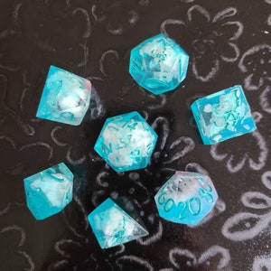 Puede incluir: Conjunto de siete dados azul turquesa con números blancos y motas blancas en todo el dado. Los dados están sobre una superficie negra con un patrón floral.