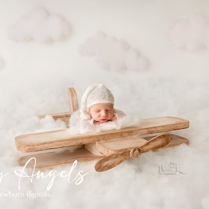 May include: A newborn baby wearing a white knitted hat is lying on a wooden airplane prop, surrounded by white fluffy clouds. The text "Tiny Angels newborn digitals" is visible at the bottom of the image.