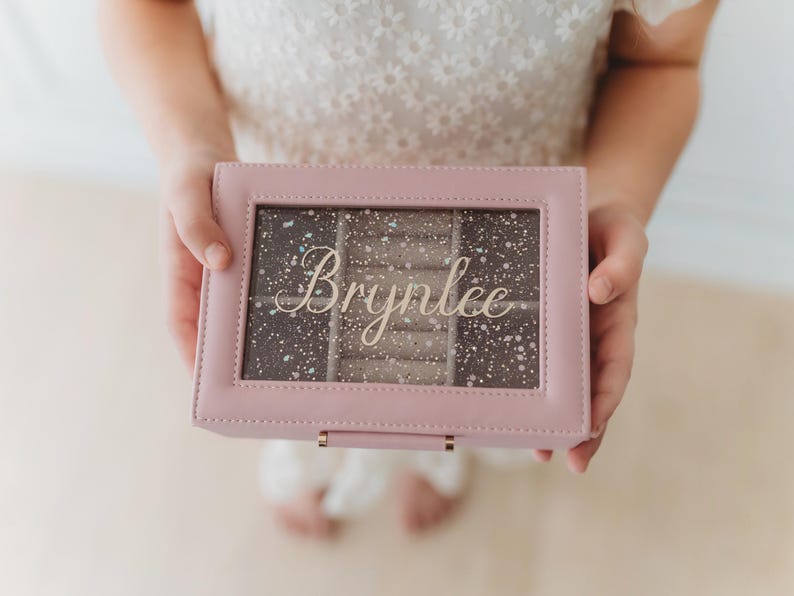 K&ouml;nnte beinhalten: Eine rosafarbene Schmuckschatulle mit einem durchsichtigen Deckel und einem funkelnden, glitzernden Innenraum. Die Schachtel tr&auml;gt eine goldene Inschrift mit dem Namen "Brynnlee".