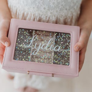 K&ouml;nnte beinhalten: Eine rosafarbene Schmuckschatulle mit einem durchsichtigen Deckel und einem glitzernden Hintergrund. Die Schachtel hat einen goldenen Verschluss und der Name "Lydia" ist auf dem Deckel geschrieben.