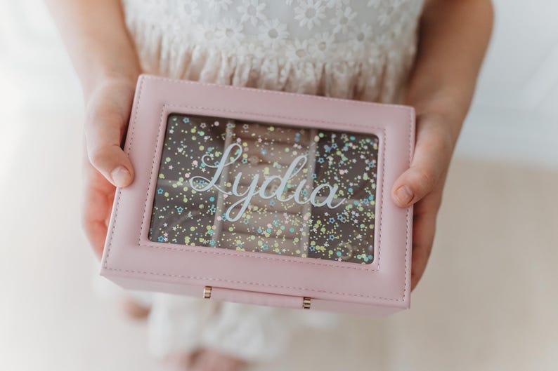 K&ouml;nnte beinhalten: Eine rosafarbene Schmuckschatulle mit einem durchsichtigen Deckel und einem glitzernden Hintergrund. Die Schachtel hat einen goldenen Verschluss und der Name "Lydia" ist auf dem Deckel geschrieben.