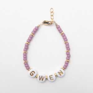 May include: A pink beaded bracelet with a gold clasp and white beads spelling out the name "GWEN".