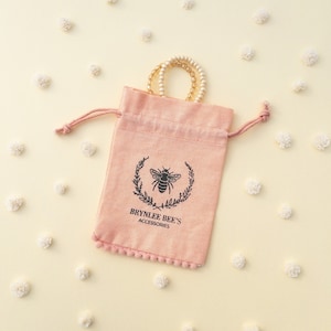 May include: A pink drawstring bag with a bee logo and the text "BRYNLEE BEE'S ACCESSORIES". The bag is filled with two gold bracelets.