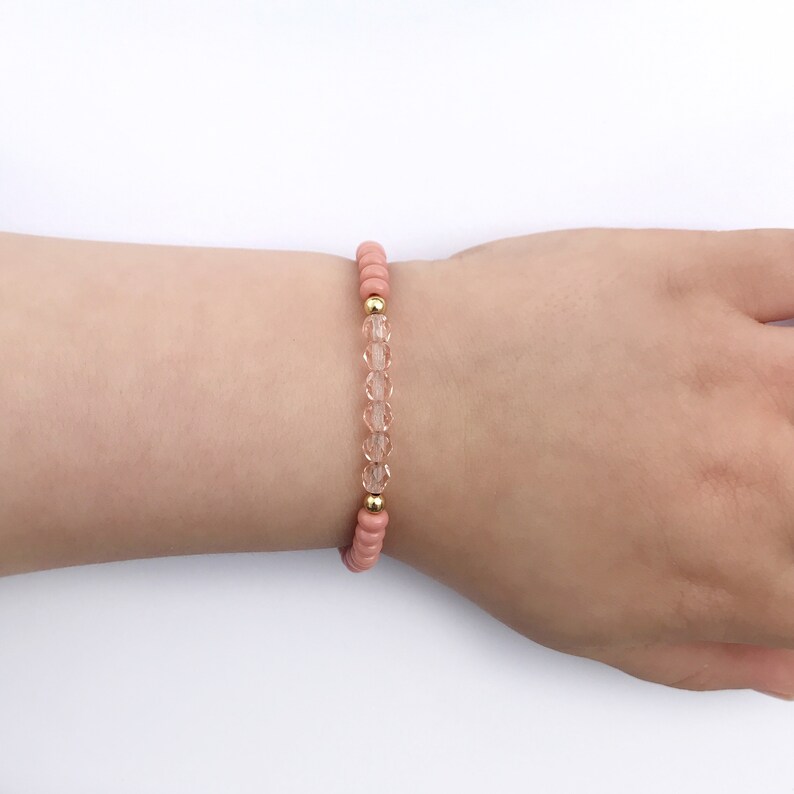 May include: A delicate bracelet with small, clear glass beads and gold accents. The bracelet is made with peach-colored beads and is worn on a wrist.