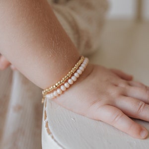 May include: A set of two bracelets for a baby or toddler. One bracelet is made of gold beads and the other is made of light pink beads.