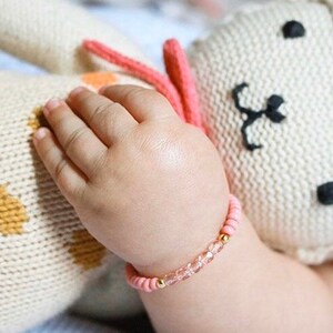 May include: A pink and gold beaded bracelet with clear crystals. The bracelet is on a baby's wrist.