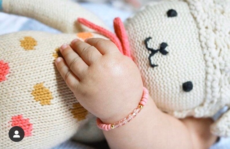 May include: A pink and gold beaded bracelet with clear crystals. The bracelet is on a baby's wrist.