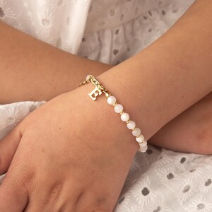 May include: A white beaded bracelet with a gold charm in the shape of the letter "E".