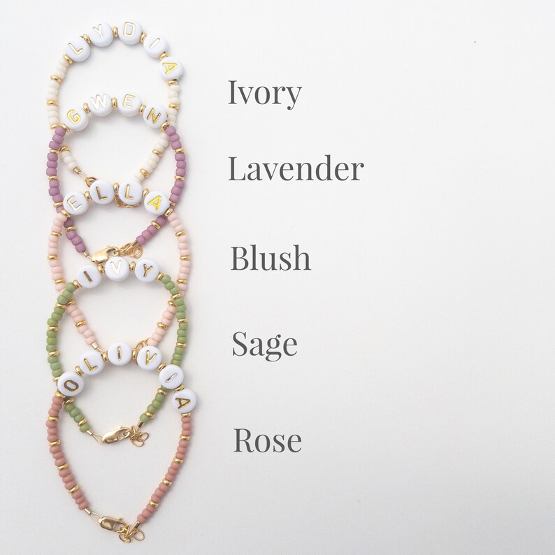 May include: Five beaded bracelets with different coloured beads and gold letter beads spelling out names. The bracelets are arranged vertically from top to bottom with the names:  Lydia, Gwen, Ella, Ivy, and Olivia. The colours of the bracelets are ivory, lavender, blush, sage, and rose.