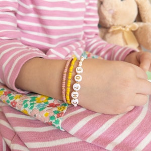 May include: A child's wrist with four bracelets. The top bracelet is white with gold letters spelling "CHLOE". The other bracelets are pink and yellow.