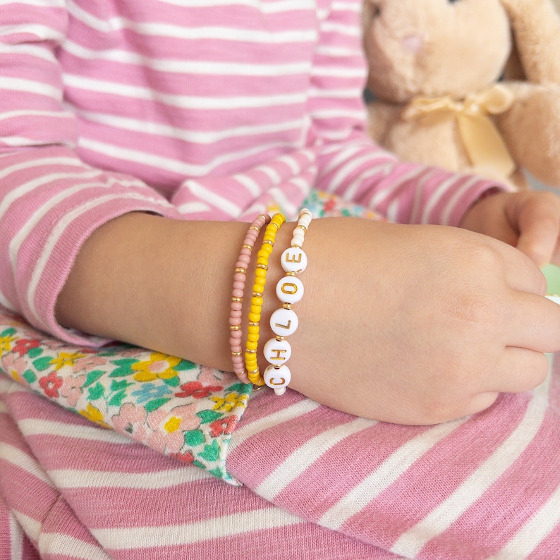 May include: A child's wrist with four bracelets. The top bracelet is white with gold letters spelling "CHLOE". The other bracelets are pink and yellow.