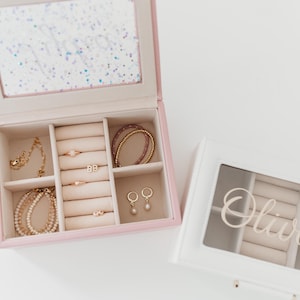 K&ouml;nnte beinhalten: Zwei Schmuckk&auml;stchen mit einem verspiegelten Innenraum und F&auml;chern f&uuml;r Ringe, Ohrringe und Armb&auml;nder. Die rosa Box hat einen glitzernden Deckel und die wei&szlig;e Box hat einen durchsichtigen Deckel mit dem Namen "Olivia" darauf.