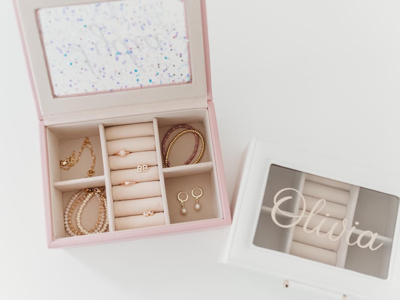K&ouml;nnte beinhalten: Zwei Schmuckk&auml;stchen mit einem verspiegelten Innenraum und F&auml;chern f&uuml;r Ringe, Ohrringe und Armb&auml;nder. Die rosa Box hat einen glitzernden Deckel und die wei&szlig;e Box hat einen durchsichtigen Deckel mit dem Namen "Olivia" darauf.