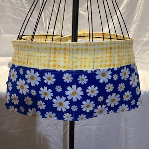 Può includere: Un grembiule in vita con una base in tessuto blu con un motivo floreale di margherite bianche e una fascia in vita a quadretti vichy gialli e bianchi. Il grembiule include lacci in tessuto sul retro ed è esposto su un manichino in filo nero.
