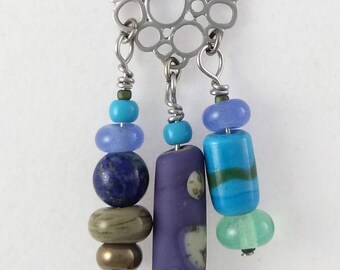 May include: A silver chain necklace with a circular pendant made of interconnected circles. The pendant has three strands of beads hanging from it. The beads are purple, blue, green, and brown.