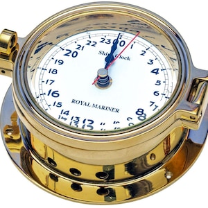 May include: A round brass ship clock with a white face featuring black numerals from 1 to 23 and the text "Ship Clock" and "ROYAL MARINER". The clock has a hinged brass frame, a glass cover, and black hour and minute hands with a red second hand.