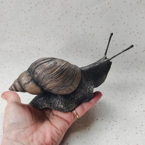 May include: A detailed sculpture of a snail with a textured, brown and tan shell and a dark gray body. The snail's antennae are long and thin. The sculpture is held in a hand, showcasing its size and detail.
