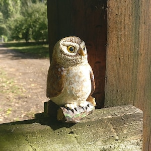 以下が含まれることがあります： 木製のフェンスの支柱の上に置かれた、茶色と白の小さなフクロウの置物。フクロウは大きく丸い目と、詳細な質感のある表面をしています。