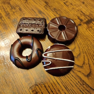 May include: Four chocolate-covered treats, including a round donut-shaped treat with a chocolate glaze and a ring of chocolate, a round chocolate treat with a white swirl design, a round chocolate treat with a starburst design, and a rectangular chocolate biscuit with the word "Bourbon" embossed on the top.