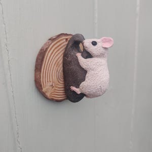 May include: A decorative wall hook featuring a light pink mouse figure climbing a dark brown, textured rock-like shape. The entire piece is mounted on a circular, natural wood slice showing visible tree rings and a light brown bark edge.