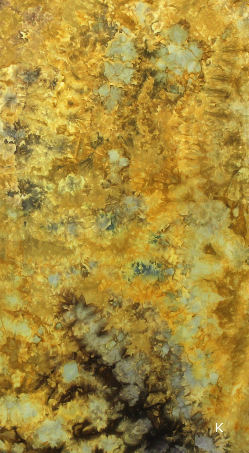 May include: A fabric with a tie-dye pattern featuring shades of golden yellow, mustard, and deep brown, with scattered light blue and white accents throughout the design. The letter K is printed in white in the bottom right corner of the fabric.